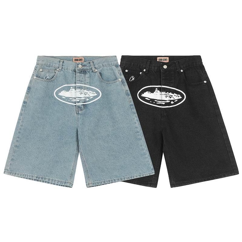 May include: Two pairs of denim shorts in light blue and black. Each pair features a white oval graphic with a central design, a button fly, belt loops, and a small embroidered logo on the right pocket. The inner waistband label reads Denim Genes.
