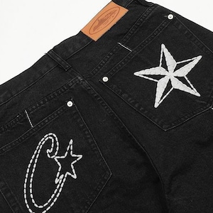 May include: Black denim jeans featuring a tan leather patch on the waistband. The back pockets are decorated with white stitched embroidery, including a large star on the right pocket and a stylized letter C with a small star on the left pocket.