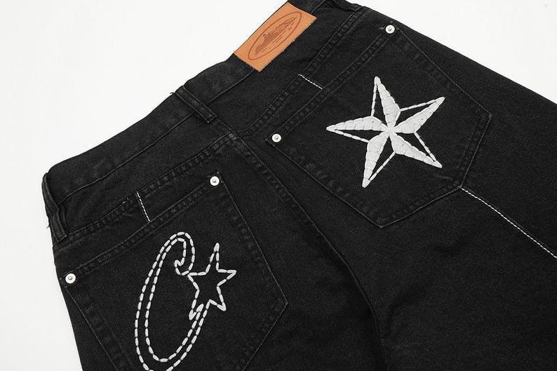 May include: Black denim jeans featuring a tan leather patch on the waistband. The back pockets are decorated with white stitched embroidery, including a large star on the right pocket and a stylized letter C with a small star on the left pocket.