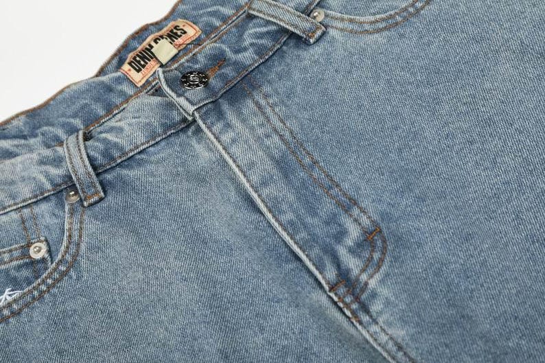 Peut inclure: Un jean en denim bleu clair dot&eacute; d'une coupe classique &agrave; cinq poches, d'une fermeture &agrave; bouton m&eacute;tallique et de passants de ceinture. Une &eacute;tiquette de marque en similicuir beige avec le texte DENIM GENES est cousue sur la ceinture int&eacute;rieure.