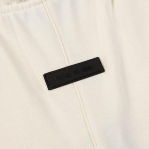 May include: An off-white fabric garment featuring a vertical seam down the center and a rectangular black rubberized patch stitched onto the front. The patch displays the embossed text FEAR OF GOD in a clean, sans-serif capital letter font.