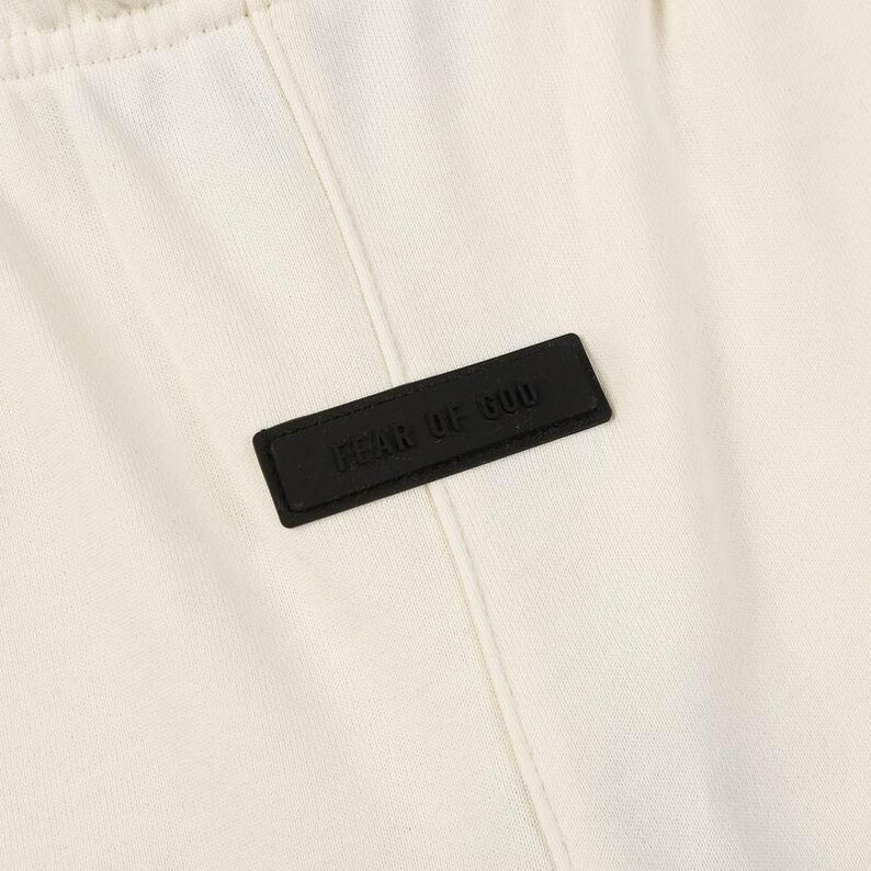 May include: An off-white fabric garment featuring a vertical seam down the center and a rectangular black rubberized patch stitched onto the front. The patch displays the embossed text FEAR OF GOD in a clean, sans-serif capital letter font.