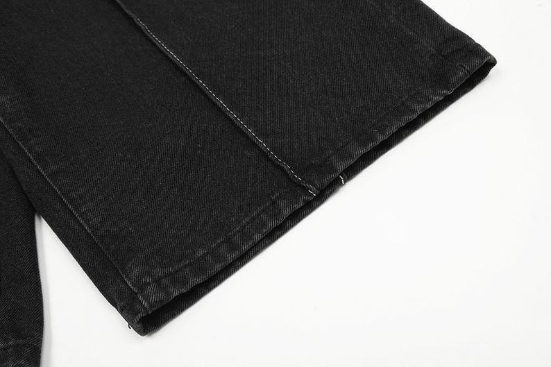 May include: A pair of black denim pants featuring a straight-leg cut and a prominent vertical seam running down the front of the leg. The fabric has a visible twill texture with clean, finished edges at the hem and a single row of white stitching.