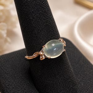 Puede incluir: Un anillo con una piedra preciosa cabujón de color verde pálido, lisa, ovalada y translúcida, engastada en una banda de metal pulido de color rosa. El engaste incluye delicados detalles metálicos en espiral que enmarcan la piedra central.