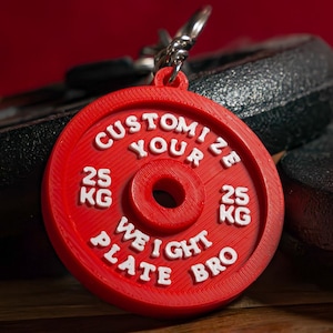 May include: A red weight plate keychain with white text that reads "Customize Your Weight Plate Bro." The keychain has a silver ring and is next to two black weight plates. Each plate is marked with "25 KG."