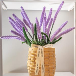 May include: A decorative arrangement of lavender-colored flowers in a woven basket. The basket is tied with a ribbon that reads "JUST FOR YOU". The flowers have green stems and are arranged to stand upright.