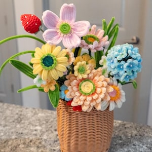 May include: A woven basket filled with a colorful assortment of crocheted flowers. The arrangement includes yellow, orange, pink, and blue flowers, along with a red crocheted strawberry. The basket is made of light brown woven material.