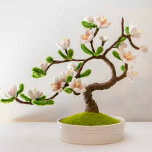 Peut inclure: Une sculpture artisanale de bonsaï avec des fleurs blanches et roses, des feuilles vertes et un tronc brun. L'arbre est dans un pot en céramique blanc rempli de mousse verte. Un élément décoratif.