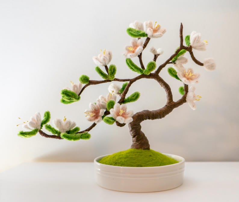 May include: A handcrafted bonsai tree sculpture with white and pink flowers, green leaves, and a brown trunk. The tree sits in a white ceramic pot filled with green moss. A decorative piece for home decor.
