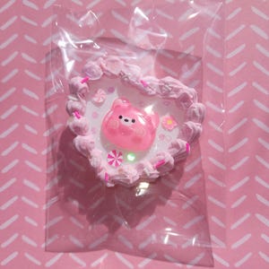 May include: A pink heart-shaped accessory featuring a clear, plastic bear at its centre. The heart is decorated with pink frosting, flowers, and a star. The accessory is packaged in a clear plastic bag.