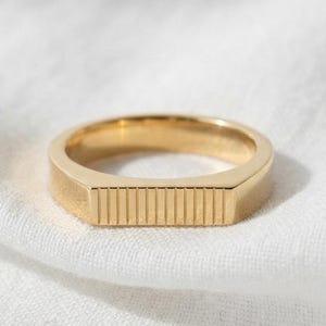 May include: A gold-toned ring featuring a flat, rectangular front section with a series of vertical, parallel engraved lines. The band is smooth and circular, maintaining a consistent width throughout its design, showcasing a polished metallic finish.