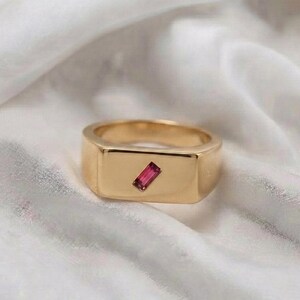 May include: A gold signet ring featuring a smooth, polished band and a flat rectangular face. A single, small, rectangular-cut pink gemstone is set diagonally in the center of the ring face, creating a clean and simple design for this piece of jewelry.