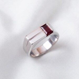 May include: A polished silver-toned ring featuring a rectangular, deep red gemstone set into one side of the band. The band has a smooth, flat surface with a distinct vertical groove running parallel to the gemstone setting, creating a clean design.