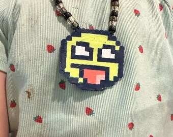 May include: A handmade necklace featuring a pixelated yellow and blue face pendant with a red tongue. The necklace has a beaded chain with black and clear beads. The person is wearing a light blue shirt with a strawberry pattern.