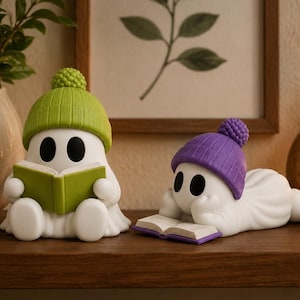 May include: Two white ghost figurines. One ghost sits upright wearing a lime green knit hat with a pom-pom, holding an open green book. The second ghost lies on its stomach wearing a purple knit hat with a pom-pom, reading an open purple book.