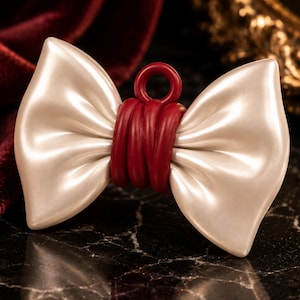 May include: A decorative bow charm featuring a smooth, glossy off-white finish on the wings and a deep red center knot with a small circular loop at the top for attachment. The bow has a structured, molded appearance with soft, curved edges.