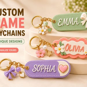 Puede incluir: Tres llaveros personalizados con anillas y cadenas doradas con dijes. Un llavero ovalado verde dice EMMA con un lazo blanco. Un llavero festoneado color melocotón dice OLIVIA con flores. Un llavero ovalado morado muestra el nombre SOPHIA con un corazón.