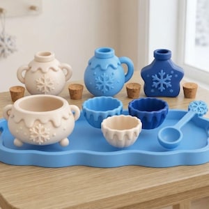 Puede incluir: Una bandeja de servicio azul contiene un conjunto de utensilios de cocina de silicona que incluye una olla beige con tapa, un frasco azul con tapa, un frasco azul en forma de estrella, tres cuencos pequeños en beige y azul, una cuchara azul y cuatro tapones de corcho.