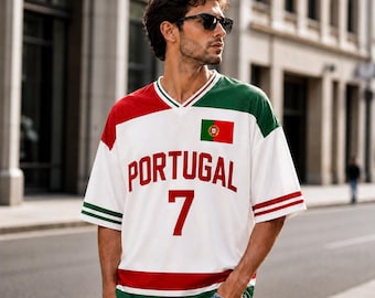 Puede incluir: Una camiseta blanca de manga corta con cuello en V y detalles en rojo y verde. El frente muestra la palabra PORTUGAL en letras rojas sobre el número 7, con un pequeño emblema de la bandera de Portugal en la parte superior izquierda.