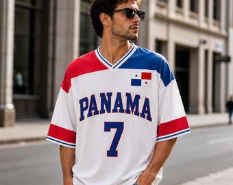 Puede incluir: Una camiseta de malla blanca, roja y azul con la palabra PANAMA en letras grandes azules en el pecho, el número 7 en azul debajo y un pequeño parche de la bandera de Panamá en el lado izquierdo, con cuello en V y mangas cortas.