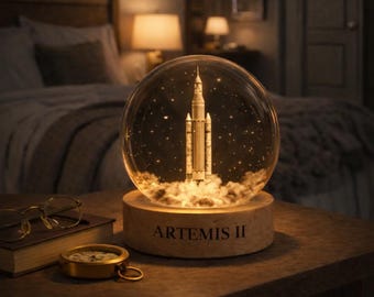 Puede incluir: Un globo de nieve decorativo con un cohete despegando, con el texto "ARTEMIS II" en la base de madera. El cohete es blanco y beige, sobre un fondo estrellado. El globo está iluminado, creando un brillo cálido y acogedor.