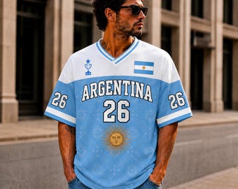 Puede incluir: Una camiseta de cuello en V azul claro y blanca con el texto ARGENTINA, el número 26, un pequeño icono de trofeo, la bandera argentina y un emblema solar central. Las mangas tienen rayas blancas y el número 26 en cada lado.