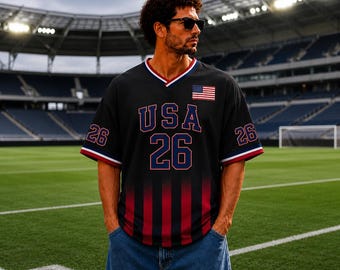 Puede incluir: Una camiseta negra con cuello en V que presenta el texto USA y el número 26 en azul oscuro con bordes rojos. La prenda incluye un pequeño parche de la bandera estadounidense en el pecho, el número 26 en las mangas y rayas verticales rojas.