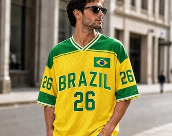 Puede incluir: Una camiseta de manga corta amarilla y verde con la palabra BRAZIL en letras verdes grandes, el número 26 en la parte delantera y las mangas, y un pequeño emblema de la bandera brasileña en el pecho. La tela tiene un sutil patrón geométrico.