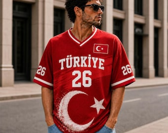 Puede incluir: Una camiseta roja de cuello en V con el texto TÜRKIYE, el número 26 y un gran gráfico blanco de una luna creciente y una estrella. La prenda incluye un pequeño emblema de la bandera turca en el pecho y el número 26 en las mangas con detalles blancos.