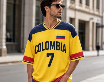 Puede incluir: Una camiseta de cuello en V amarilla con hombros y detalles en las mangas de color azul marino, con la palabra COLOMBIA en letras azul marino, el número 7 en azul marino y una pequeña bandera de Colombia en el pecho, con vaqueros azul claro.