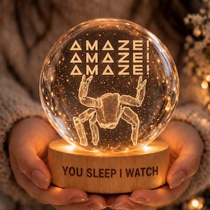 May include: A glowing glass sphere with a spider design and the word "AMAZE!" repeated three times. The sphere sits on a wooden base with the text "YOU SLEEP I WATCH". The light is warm and inviting.