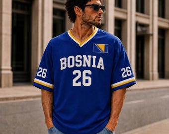 Puede incluir: Una camiseta azul de manga corta con cuello en V que presenta el texto BOSNIA y el número 26 en blanco en la parte frontal. Las mangas tienen rayas amarillas y blancas, y la bandera de Bosnia aparece en el pecho izquierdo. Se usa con jeans azules.