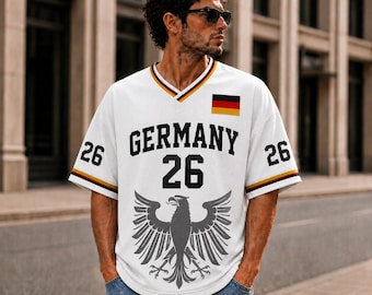 Puede incluir: Una camiseta blanca de cuello en V con la palabra GERMANY en letras negras, el número 26 en negro en el pecho y las mangas, una bandera alemana y un gran emblema de águila gris en la parte delantera con ribetes negros, rojos y dorados.