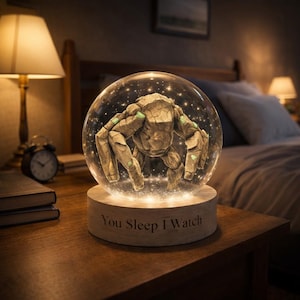 Peut inclure: Un globe de neige décoratif avec une créature en pierre à l'intérieur d'une sphère en verre transparent. La base du globe est de couleur claire et porte l'inscription "You Sleep I Watch". Le globe est éclairé, créant une lueur chaleureuse.