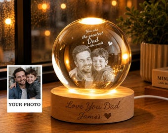 Puede incluir: Una esfera de cristal personalizada con una foto de un padre y un niño en su interior. La esfera se apoya en una base de madera con el texto "Love You Dad James" y un corazón. La esfera también tiene el texto "You are the greatest Dad in the world."