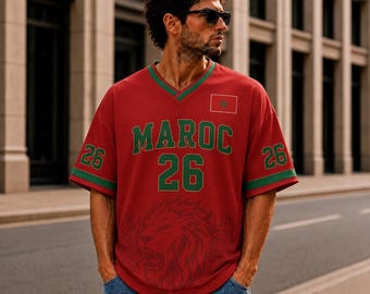 Puede incluir: Una camiseta roja de cuello en V con el texto MAROC y el número 26 en verde. La prenda incluye un pequeño gráfico de la bandera de Marruecos, ribetes verdes en las mangas y una gran ilustración sutil de una cabeza de león en el frente.