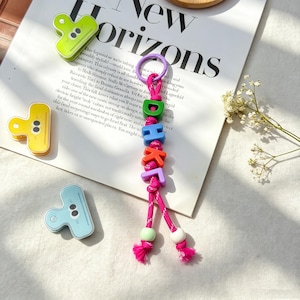 May include: A colorful keychain with the letters "DOKI" spelled out in vibrant, block-style beads. The keychain features a pink braided cord, a purple ring, and two decorative beads. The background includes paper clips and a magazine.