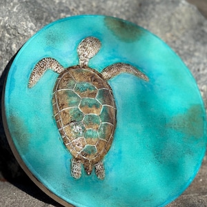 May include: A circular, turquoise decorative item with a detailed, textured sea turtle. The turtle's shell is brown and teal, with its flippers and head extended. The piece has a light brown border.