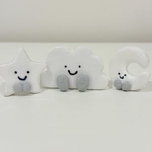 May include: Three small white ceramic figures shaped like a star, a cloud, and a crescent moon. Each figure features a glossy finish, a hand-painted black smiling face, and two small gray rounded feet positioned at the base of each object.