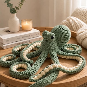 Könnte beinhalten: Ein salbeigrünes, gehäkeltes Oktopus-Stofftier mit einem großen, abgerundeten Kopf, zwei markanten schwarzen Augen und acht langen Tentakeln mit einer cremefarbenen, strukturierten Unterseite. Das Garn ist durchgehend fest verarbeitet.