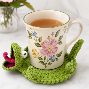 Könnte beinhalten: Ein Keramikbecher mit Blumenmuster in Rosa, Gelb und Blau, der auf einem grünen Häkeluntersetzer in Form eines Frosches mit weit geöffnetem Maul, weißen Augen, einer rosa Zunge und einem kleinen Schwanz an der Rückseite steht.