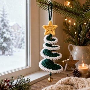 Op de afbeelding: Een spiraalvormige kerstboomversiering gehaakt van donkergroen garen met een witte rand langs de zijkanten. De versiering heeft een gele gehaakte ster aan de bovenkant en een klein goudkleurig metalen belletje dat aan de onderkant hangt.