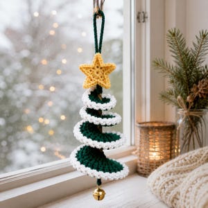 Op de afbeelding: Een spiraalvormige kerstboomhanger van haakwerk in donkergroen met een witte rand. Het ornament is voorzien van een gele gehaakte ster aan de bovenkant, een donkergroene ophanglus en een klein goudkleurig metalen belletje aan de onderkant.