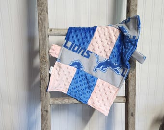 May include: A baby blanket featuring a patchwork design with blue, pink, and gray panels. The blanket displays the word "LIONS" and a lion logo. The blanket is made of soft, textured fabric and is displayed on a wooden ladder.