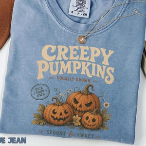 Puede incluir: Una camiseta azul de manga corta con un estampado gráfico de tres calabazas y el texto "CREEPY PUMPKINS LOCALLY GROWN PICK YOUR OWN SPOOKY & SWEET". La etiqueta de la marca Comfort Colors en el cuello interior muestra la talla S.