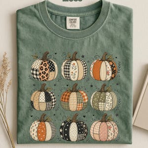 Puede incluir: Una camiseta verde musgo con un estampado gráfico de nueve calabazas dispuestas en cuadrícula. Cada calabaza muestra un patrón de patchwork único que incluye estampados de leopardo, cuadros, flores y lunares en tonos naranja, negro y blanco.