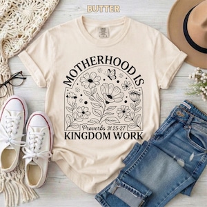 Puede incluir: Camiseta color crema con el texto "MOTHERHOOD IS KINGDOM WORK" en un diseño floral negro. La camiseta se combina con vaqueros azules, zapatillas blancas y un sombrero beige. También es visible el texto "Proverbios 31:25-27".