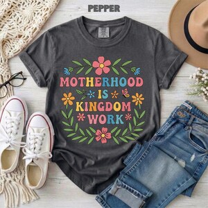 Puede incluir: Camiseta gris oscuro con el texto "MOTHERHOOD IS KINGDOM WORK" en letras coloridas rodeadas de flores y hojas. La camiseta se combina con vaqueros azules y zapatillas blancas.