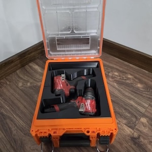 May include: An open orange tool case with a clear lid, containing two red and black power tools. The case is on a wooden floor. The tools are neatly arranged in black foam inserts.