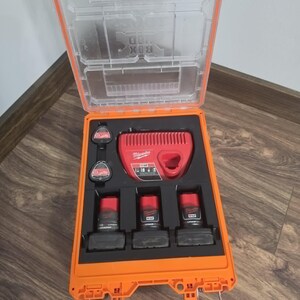 May include: An orange Milwaukee Packout tool case with a clear lid, containing a red M12 battery charger and three M12 lithium-ion batteries fitted into custom black foam inserts. The case features a sturdy handle and metal latches for secure storage.